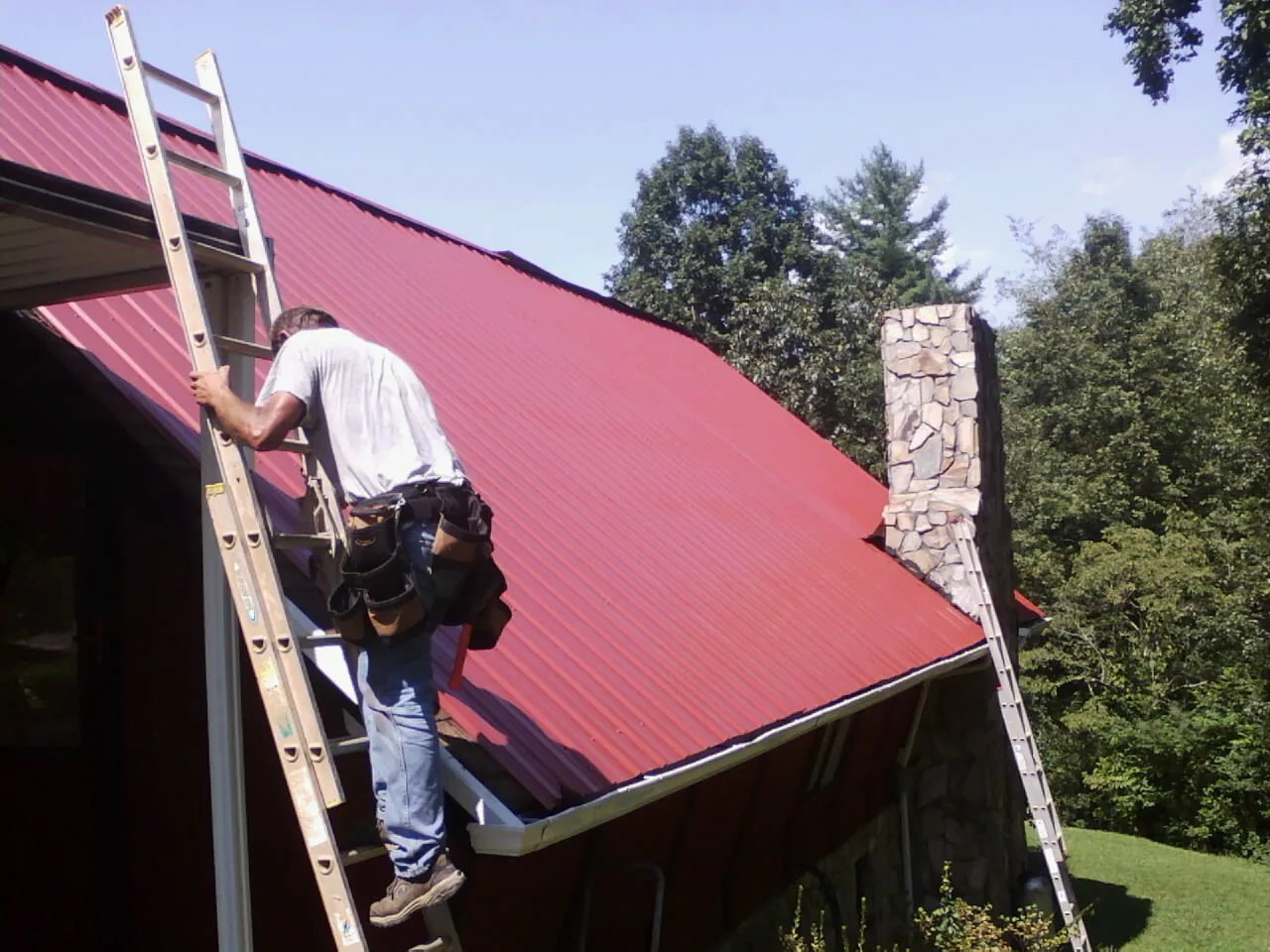 Roofer on a ladder beside a red metal roof and stone chimney — Soffit & Fascia Repair by Roofing Brothers in Glen Wild